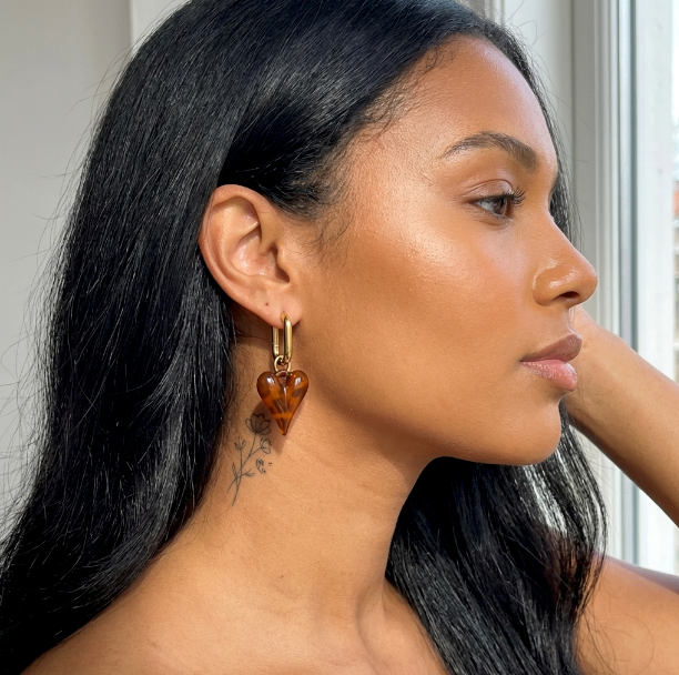 Frau trägt Safari Heart Ohrringe: goldene Hoop-Ohrringe mit braunem herzförmigen Anhänger.